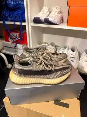 Yeezy Boost 350 V2 "Zebra" sneakers - Säljer ett par Yeezy Boost 350 V2 sneakers i färgen "Zebra" med beige, bruna och grå toner. Skorna har en stickad ovandel i Primeknit, ikonisk rand längs sidan och en tjock, ribbad sula i ljusgul ton. Perfekta för dig som gillar streetwear och vill sticka ut. Jag har lagt in stödilägg i skorna