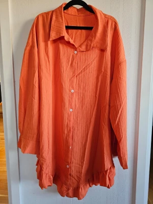 Oversized orange skjorta med volang - Säljer en oversized orange skjorta med långa ärmar och volangdetalj längst ner. Skjortan har knappar framtill och en klassisk krage. Tillverkad i ett lätt, strukturerat tyg som känns luftigt och skönt. Storlek EU52/3XL