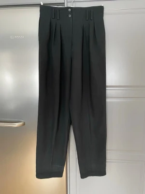 Svarta vida kostymbyxor med hög midja strl 40 - Säljer ett par svarta kostymbyxor med hög midja och vida ben. Byxorna har dubbla frontveck, breda hällor för bälte och stängs med två knappar och dragkedja. Klassisk och stilren modell som passar till många olika looks.