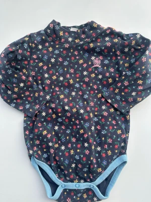 Mörkblå blommig body med blå kant - Söt mörkblå body med små färgglada blommor i rött, vitt, gult och blått. Ljusblå kant runt benen och tryckknappar i grenen. Långärmad modell med liten volangkrage. Perfekt för dig som gillar färgglada och lekfulla plagg.tvättpåverkan finns