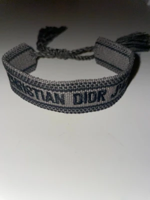 Grått Christian Dior J'Adior armband - Snyggt grått tygarmband från Christian Dior med texten 'J'Adior' broderad i svart. Armbandet har justerbar knytning med tofsar i ändarna och en stilren, trendig look som passar perfekt till din outfit.