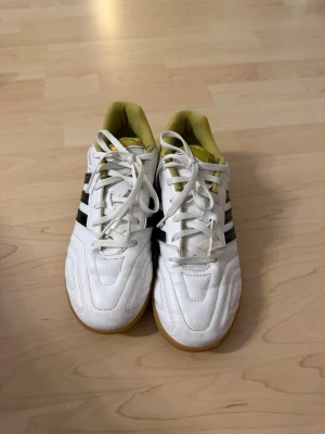Vita Adidas fotbollsskor med guld - Säljer ett par klassiska vita Adidas fotbollsskor med svarta ränder och guldfärgade detaljer vid hälen och insidan. Skorna har snörning, låg profil och gummisula som passar perfekt för inomhusspel. Tillverkade i syntetmaterial och designade för bra grepp och komfort.