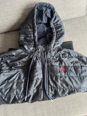 Svart pufferjacka från Polo Ralph Lauren - Snygg svart pufferjacka från Poloh ralphi talphi med huva och dragkedja framtill. Jackan har quiltad framsida i polyester och mjuka ärmar i sweatshirtmaterial. Röd broderad logga på bröstet och praktiska fickor på sidorna.