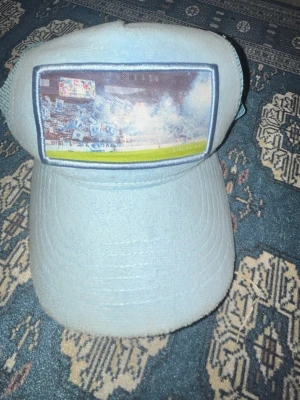 Ljusblå truckerkeps med fototryck - Säljer en ljusblå truckerkeps med mesh på sidorna och baksidan för extra ventilation. Framtill finns ett fototryck av en fotbollsläktare med tifor och rök. Justerbar storlek baktill. Perfekt för dig som gillar sportig och unik stil.