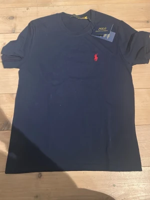 Mörkblå t-shirt från Polo Ralph Lauren - Klassisk mörkblå t-shirt från Polo Ralph Lauren med rund halsringning och ikoniska röda logobrodyr på bröstet. Tillverkad i mjuk bomull för en skön känsla och perfekt passform. Enkel och stilren design som funkar till allt.