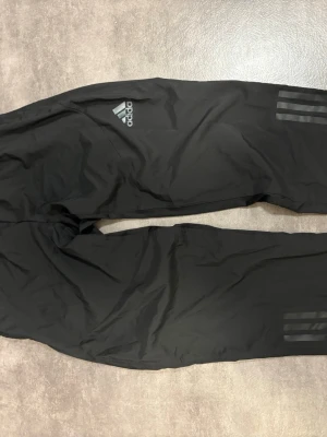 Svarta träningsbyxor från Adidas - Säljer ett par svarta träningsbyxor från Adidas med klassiska tre ränder på benen och logga på låret. Byxorna har resår i midjan och är tillverkade i ett lätt och smidigt material, perfekta för träning eller chill. Passar dig som gillar sportig stil.