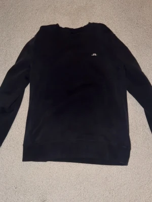Svart sweatshirt från J.Lindeberg - Snygg svart sweatshirt från J.Lindeberg med diskret logga broderad på bröstet. Tröjan har rund halsringning och ribbade muddar vid ärmslut och nederkant. Perfekt basplagg i mjukt bomullsmaterial.