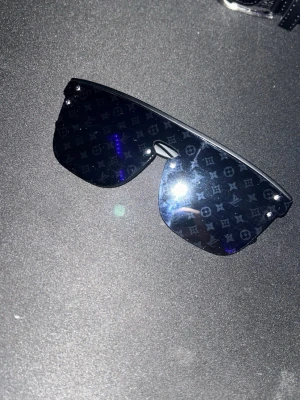 Louis Vuitton brillor - Säljer ett par svarta shield-solglasögon från Louis Vuitton med monogrammönster på glasen. Bågarna är mattsvarta och har Louis Vuitton-loggan på sidan. Glasen är stora och täcker mycket av ansiktet för en riktigt statement-look.