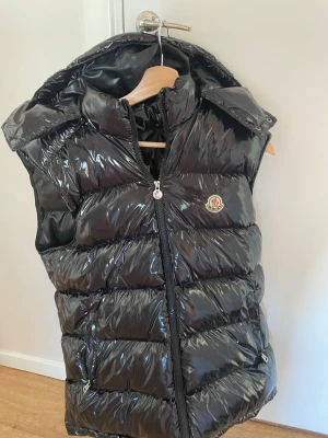 Mocler väst - Snygg svart dunväst från Moncler med glansigt yttermaterial, dragkedja framtill och två fickor med dragkedja. Västen har hög krage och ikonisk Moncler-logga på bröstet. Perfekt för lager-på-lager och streetstyle. Storlek XL