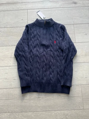 Mörkblå kabelstickad tröja Polo Ralph Lauren - Mörkblå kabelstickad tröja från Polo Ralph Lauren med halv dragkedja och hög krage. Tröjan har ett rött broderat logomärke på bröstet och ribbade muddar vid ärmslut och nederkant. Perfekt för lager på lager och en klassisk look.