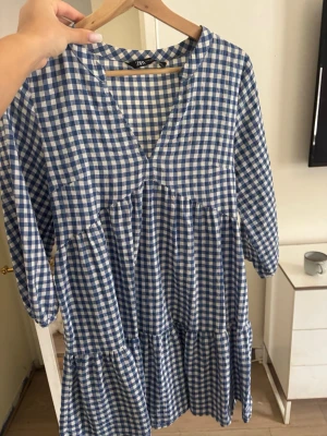 Blårutig klänning från Zara - Supersöt blårutig klänning från Zara med v-ringning och trekvartsärm. Klänningen har en luftig passform och volangdetaljer i kjolen. Perfekt för vår och sommar när du vill ha en avslappnad men trendig look.