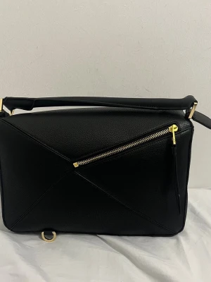 Svart Puzzle Bag från Loewe - Säljer en svart Loewe Puzzle Bag i läder med geometrisk design och guldfärgade metalldetaljer. Väskan har ett snedställt blixtlås framtill och präglad Loewe-logga i hörnet. Perfekt axelrem för att bära crossbody eller på axeln.