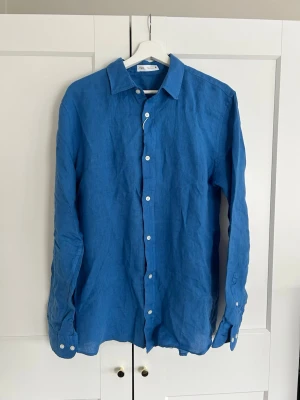 Blå linneskjorta från Zara - Snygg blå linne skjorta från Zara. Klassisk krage, vita knappar framtill och långa ärmar med knapp vid manschetten. Skjortan har en rak passform och är superlätt att matcha.
