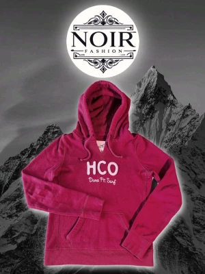 Vinröd hoodie från Hollister  - Snygg vinröd hoodie från Hollister med broderad vit text 'HCO Dana Pt. Surf' på bröstet. Klassisk känguruficka framtill och stor huva. Perfekt för en avslappnad och trendig look. Mjuk bomullsblandning som känns skön mot huden.