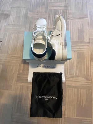 Philippe Model vita sneakers - Stilrena vita sneakers från Philippe Model med detaljer i grått och svart. Skorna har en låg silhuett, vita skosnören och en robust sula. Tillverkade i läder med märkets logotyp på sidan. Perfekta för en clean och modern look.