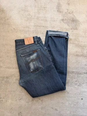STEADY EDDIE DRY GREYTONE SELVAGE 33/32 - STEADY EDDIE DRY GREYTONE SELVAGE. Säljer ett par klassiska blå jeans från Nudie Jeans med snyggt slitna detaljer på bakfickan och en läderpatch i midjan. Jeansen har rak passform och är gjorda i mjukt denimtyg. Perfekta för en avslappnad och trendig look.