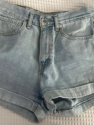 Ljusblå jeansshorts från Levi's - Ljusblå jeansshorts från Levi's med klassisk femficksdesign, uppvikta benslut och gylf med dragkedja. Shorts i bomull med Levi's ikoniska läderpatch och röd logotagg bak. Perfekta för varma dagar och enkel att matcha med allt.