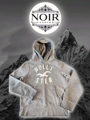 Grå hoodie från Hollister - Snygg grå hoodie från Hollister med stor vit logga och text på bröstet. Klassisk huva med dragsko och magficka framtill. Tillverkad i mjukt sweatshirtmaterial som är perfekt för chill dagar. Ribbade muddar vid ärmslut och nederkant.