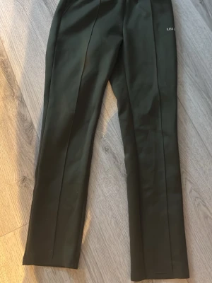 Mörkgröna mjukisbyxor LES DEUX - Mörkgröna mjukisbyxor från LES DEUX med raka ben och resår i midjan. Byxorna har diskreta sömmar längs benen och en liten logga på höften. Perfekta för en chill och avslappnad stil.
