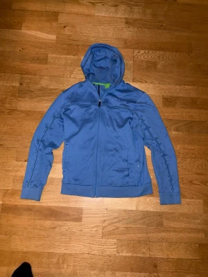Blå hoodie från BOSS med dragkedja - Snygg blå hoodie från BOSS med hel dragkedja framtill och huva med BOSS-logga längs kanten. Tillverkad i mjukt bomullsmaterial och har ribbade muddar vid ärmslut och nederkant. Diskret BOSS-tryck på bröstet och praktiska fickor framtill.