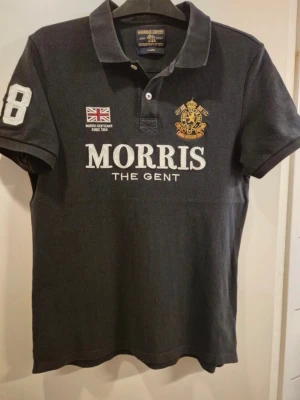 Svart pikétröja från Morris - Snygg svart pikétröja från Morris med broderad logga, brittisk flagga och siffran 68 på ärmen. Klassisk krage, kortärmad och med knappar framtill. Perfekt för en stilren och sportig look.