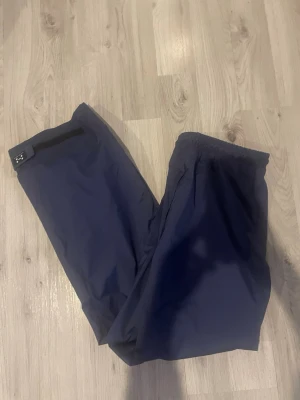 Mörkblå täckbyxor från haglöfs - Säljer ett par mörkblå täckbyxor från Haglöfs med justerbar kardborre i midjan. Byxorna har en enkel och clean design, perfekta för kalla dagar. Materialet är slitstarkt och vindtätt, vilket gör dem grymma för vinterns alla äventyr.