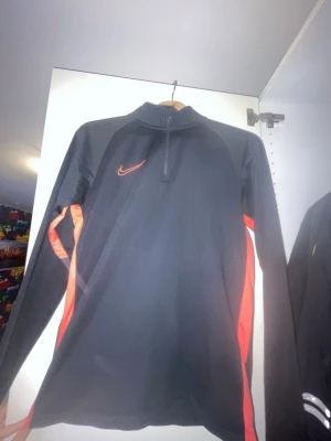 Svart träningsjacka från Nike - Snygg svart träningsjacka från Nike med röda detaljer längs sidorna och röd Nike-logga på bröstet. Jackan har halv dragkedja framtill och lång ärm, perfekt för löpning eller gym. Tillverkad i ett lätt och funktionellt material som andas.