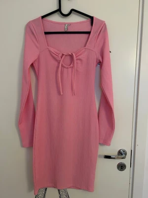 Rosa ribbad klänning med snörning från NLY Trend - Rosa ribbad klänning med lång ärm och fyrkantig ringning från NLY Trend i storlek XS. Klänningen har en snygg snörningsdetalj framtill och är i en kort modell. Endast använd en gång. 