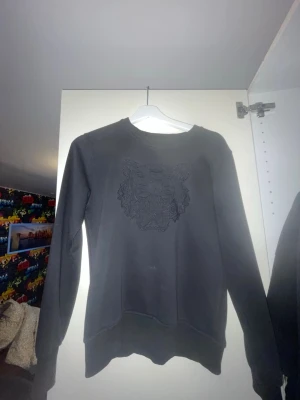 Svart sweatshirt från Kenzo - Säljer en svart sweatshirt från Kenzo med det ikoniska tigermotivet broderat på bröstet. Tröjan har rund halsringning, långa ärmar och ribbade muddar. Tillverkad i mjukt bomullsmaterial som är skönt att bära.