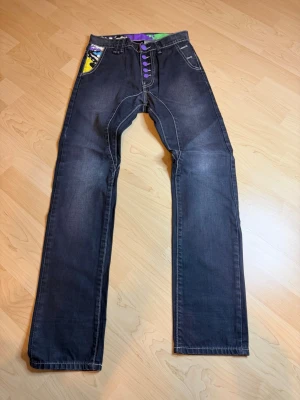 Svarta jeans med färgglada detaljer - Unika svarta jeans med raka ben och färgglada detaljer vid fickorna och knäppningen. Jeansen har lila knappar framtill och en stor bakficka med grafiskt tryck i neonfärger. Perfekta för dig som vill sticka ut med din stil.