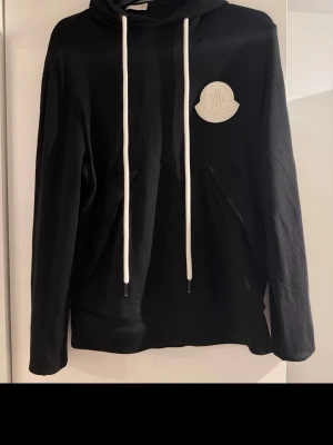 Svart Moncler hoodie med logga - Snygg svart hoodie från Moncler med stor vit logga på bröstet och vita dragsnören i huvan. Ärmen har Moncler-tryck i vitt längs hela sidan. Klassisk känguruficka framtill och relaxed fit. Perfekt för dig som gillar streetwear och exklusiva märken.