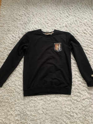 Svart sweatshirt från Burberry - Snygg svart sweatshirt från Burberry med klassisk rutmönstrad patch och broderad logga på bröstet. Tröjan har rund halsringning och långa ärmar. Perfekt för dig som gillar stilrena och exklusiva plagg. Står l men sitter som s 