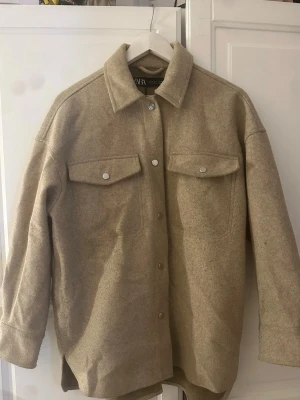 Beige kappa från Zara - Stilren beige kappa från Zara med klassisk krage och tryckknappar framtill. Två stora bröstfickor med lock och knappdetaljer. Kappan är gjord i ett mjukt, ull-liknande material och har en avslappnad passform som passar perfekt till lager på lager.