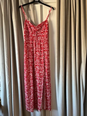 Röd blommig långklänning med smala band - Snygg röd långklänning med vita blommor och smala axelband. Klänningen har en lätt v-ringad urringning och ett feminint fall. Perfekt för varma dagar och sommarens alla tillfällen. Materialet känns mjukt och luftigt. Använd fåtal gånger.