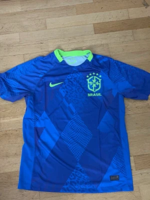Blå Brasilien Nike fotbollströja - Snygg blå fotbollströja från Nike med Brasilien-logga och gula detaljer. Tröjan har korta ärmar, rund hals med limegrön kant och ett mönster i olika blå nyanser. Perfekt för dig som älskar fotboll och vill sticka ut på planen.