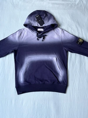 Blå hoodie från Stone Island - Snygg blå hoodie från Stone Island med cool gradient-effekt från mörkblått till ljusare blått. Klassisk känguruficka framtill, huva med dragsko och ikonisk Stone Island-patch på ärmen. Perfekt för dig som vill ha en streetwear-look med premiumkänsla.