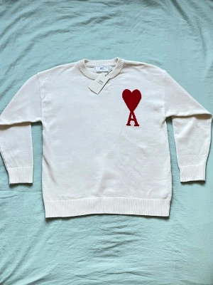 Vit stickad tröja från Ami Paris - Säljer en vit stickad tröja från Ami Paris med deras ikoniska röda hjärta och bokstaven A broderat på bröstet. Tröjan har rund halsringning, ribbade muddar och är tillverkad i mjukt material. Perfekt för dig som gillar stilrena plagg med en twist.