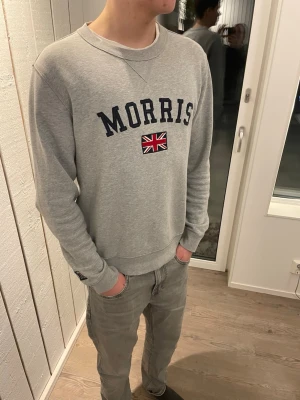 Morris tröja  - Snygg grå sweatshirt från Morris med broderad logga och brittisk flagga på bröstet. Tröjan har rund halsringning, ribbade muddar och en patch på ärmen. Perfekt för en avslappnad och stilren look.