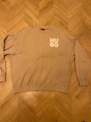 Beige sweatshirt från Hugo - Snygg beige sweatshirt från Hugo med vit logga tryckt på bröstet. Tröjan har rund halsringning, ribbade muddar vid ärmslut och nederkant samt en avslappnad passform. Tillverkad i mjukt bomullsmaterial som känns skönt mot huden. Perfekt för en casual och trendig look.