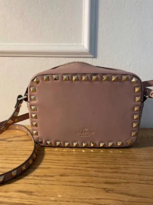Rosa axelväska Valentino med nitar - Säljer en snygg rosa axelväska från Valentino med guldfärgade nitar runt kanterna. Väskan är i läder och har en justerbar axelrem med matchande nitar. Ältighetsbevis saknar tyvärr och därav priset. 