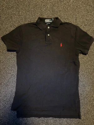 Svart pikétröja från Ralph Lauren - Klassisk svart pikétröja från Ralph Lauren med röd broderad logga på bröstet. Tröjan har korta ärmar, krage och två knappar framtill. Tillverkad i bomull för en bekväm känsla och snygg passform.