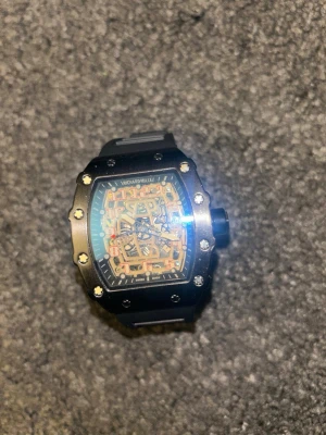 RM svart klocka - Snygg svart klocka med rektangulärt boett och detaljerad urtavla i orange och guldtoner. Klockan har ett robust svart armband i syntetmaterial och synliga skruvar på boetten för en cool och modern look.