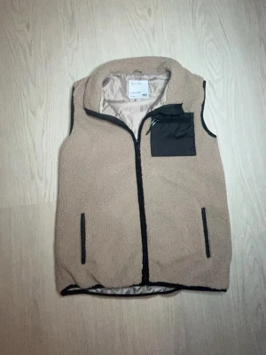 Beige fleeceväst med svarta detaljer päls - Säljer en beige fleeceväst med svarta dragkedjor och detaljer. Västen har två sidofickor och en bröstficka i svart tyg. Perfekt för lager-på-lager och snyggt stilrent!😀Bara att höra av sig vid minsta fundering!