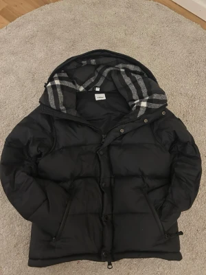 Svart pufferjacka från Burberry - Säljer en svart pufferjacka från Burberry med klassiskt rutigt foder i huvan. Jackan har dragkedja och knappar framtill, två snedställda fickor och justerbar nederkant. Perfekt för kalla dagar och riktigt snygg streetstil.