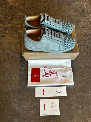 Gråa Christian Louboutin sneakers pris kan sänkas lite. - Snygga gråa sneakers från Christian Louboutin i mocka med ikoniska nitar på tån och klassisk röd sula. Skorna har lågt skaft, matchande grå skosnören och exklusiv design som sticker ut. Perfekta för dig som gillar statement-plagg och lyxiga detaljer.