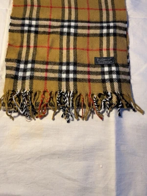Klassisk beige rutig halsduk Burberry - Snygg beige halsduk från Burberry med ikoniskt rutmönster i svart, vitt och rött. Tillverkad i 100% lammull och har fransar i ändarna. Perfekt accessoar för att lyfta din outfit med en tidlös brittisk touch.