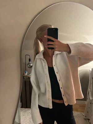 Vit stickad kofta med krage - Säljer en vit stickad kofta med stora knappar och bred krage. Koftan har en avslappnad passform, långa ärmar och ribbade muddar vid ärmslut och nederkant. Perfekt att slänga över en topp eller bara ha som den är🫶