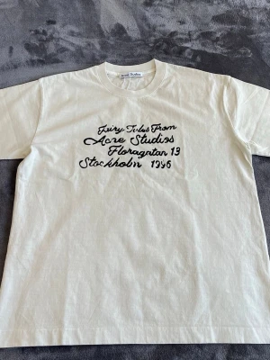 Vit t-shirt från Acne Studios - Vit t-shirt från Acne Studios med svart text på bröstet där det står 'Fairy Tales From Acne Studios Floragatan 13 Stockholm 1996'. Klassisk passform och rund halsringning. Perfekt för dig som gillar stilrena plagg med en twist.
