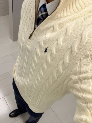 Vit kabelstickad tröja Polo Ralph Lauren - Kabelstickad vit tröja från Polo Ralph Lauren med halv dragkedja och broderad logga på bröstet. Tröjan har ribbade muddar och hög krage, perfekt för en klassisk och stilren look. Materialet är mjukt och bekvämt.