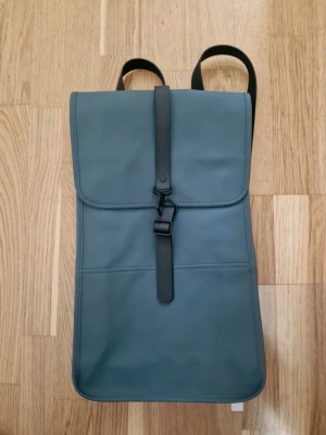 Blå minimalistisk ryggsäck från Rains - Snygg blå ryggsäck från Rains i vattenavvisande material med stilren, rektangulär form. Väskan har ett lock med magnetisk rem och metallspänne samt justerbara axelband i svart. Perfekt för dig som gillar enkel och modern design.
